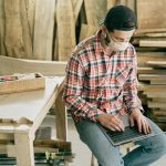 A carpenter in a workshop using a laptop, surrounded by tools and wood.